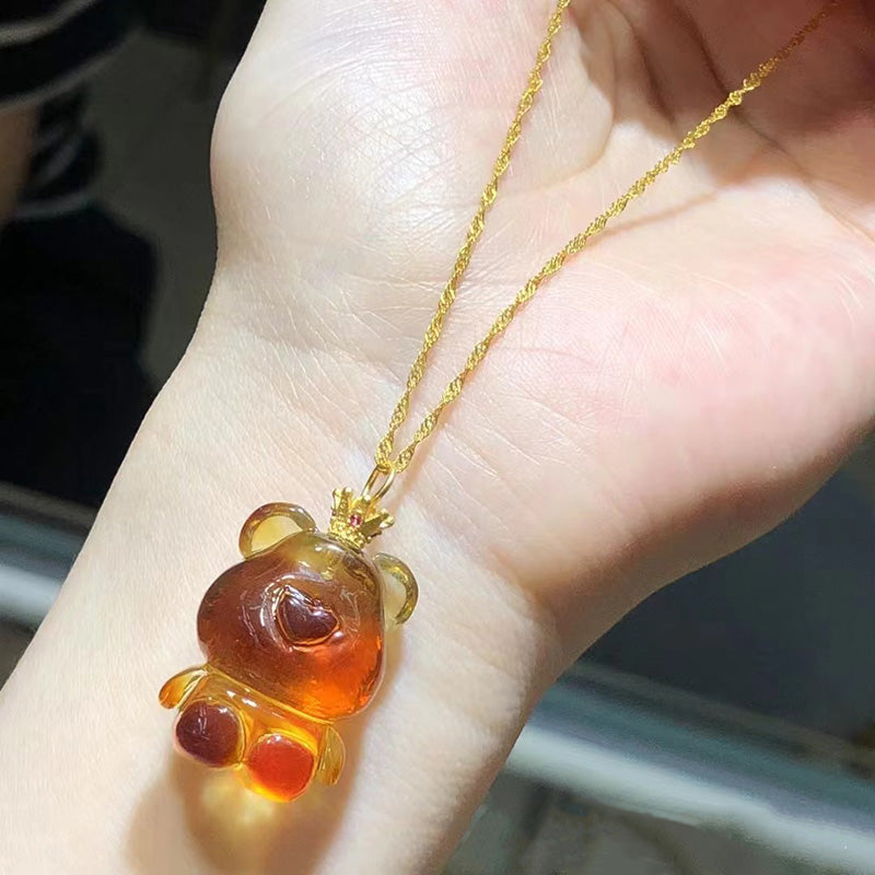 A necklace featuring a carved amber teddy bear pendant with red accents, attached to a gold - toned chain, held in a hand. 03