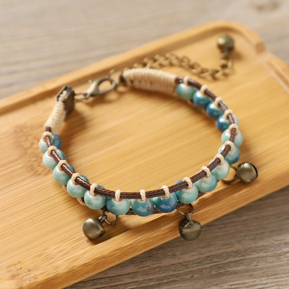 his bohemian bracelet features vibrant multicolored ceramic beads woven onto a brown leather cord, accented with small bronze bell charms. The adjustable design ensures a comfortable fit