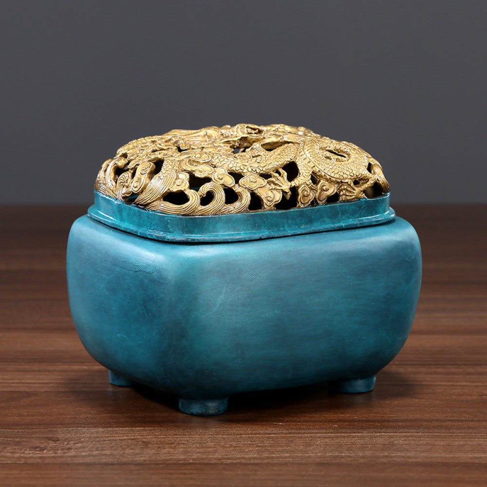 Blue-green bronze Chinese incense burner with intricately carved golden dragon pierced lid, square body, and small feet, a Feng Shui symbol of wealth and protection, displayed as a stylish home decor piece on a wooden surface against a gray background