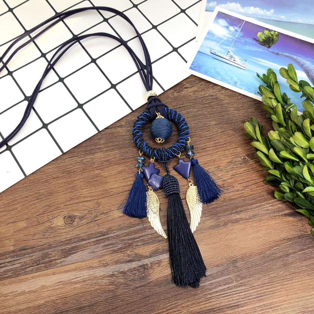 Vintage bohemian blue dreamcatcher necklace with tassel, feather, and geometric accents, displayed on a wooden surface with grid paper and greenery, ideal for boho fashion enthusiasts