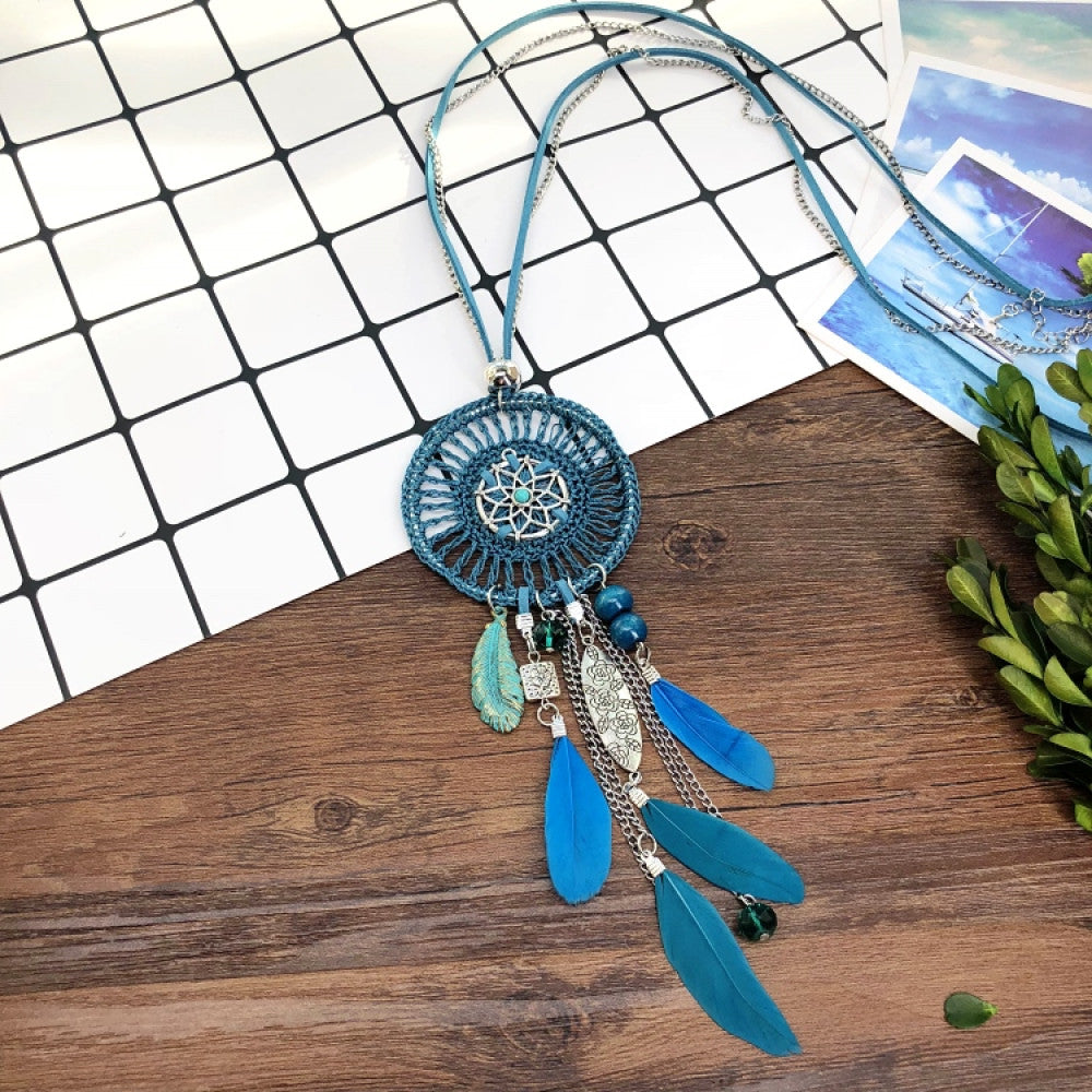 Vintage bohemian blue dreamcatcher necklace with tassel, feather, and geometric accents, displayed on a wooden surface with grid paper and greenery, ideal for boho fashion enthusiasts 03