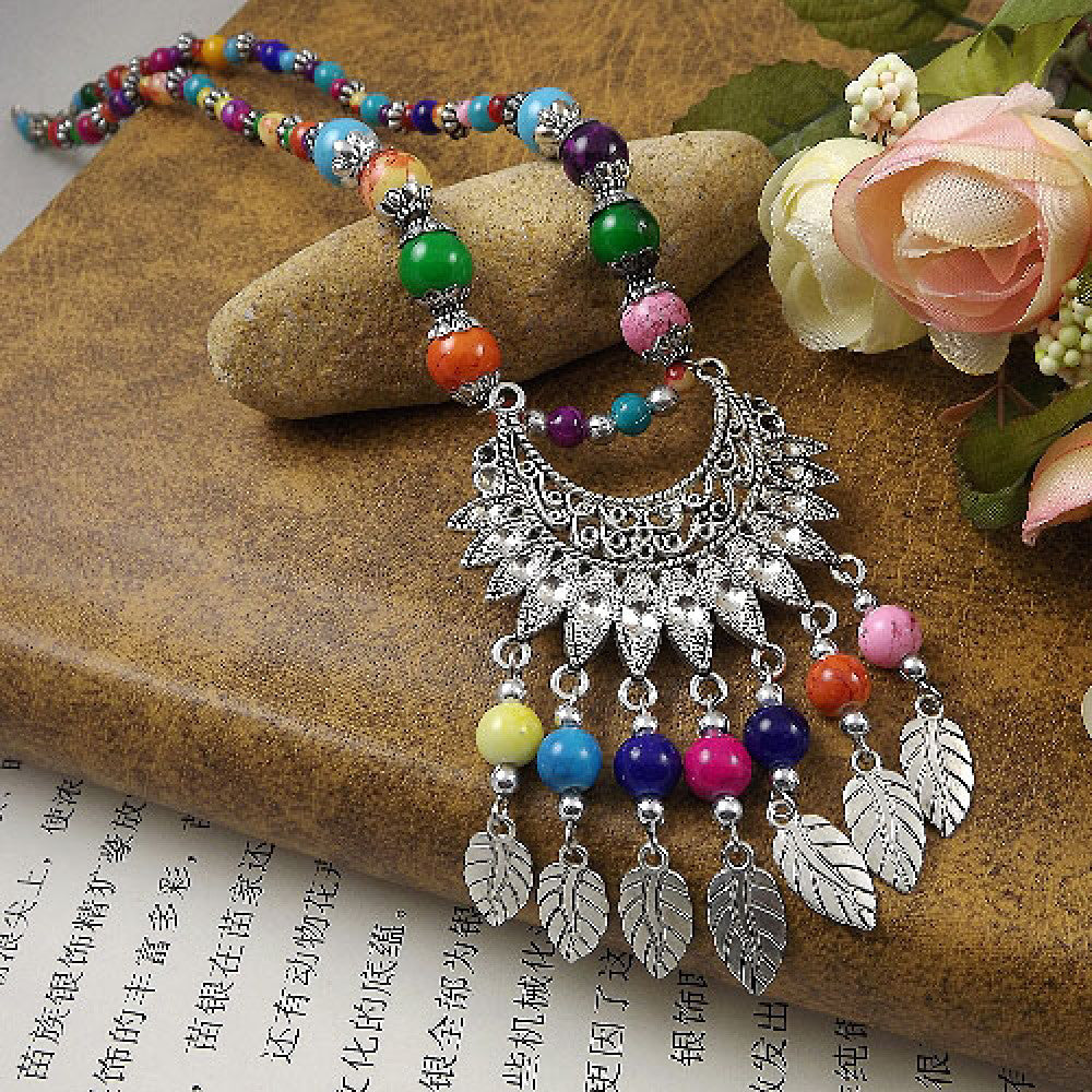 Vibrant colorful beaded necklace with intricate silver-toned leaf tassel and crescent pendant, featuring a mix of bright beads, ideal for bohemian fashion, showcased on a textured surface with floral accents