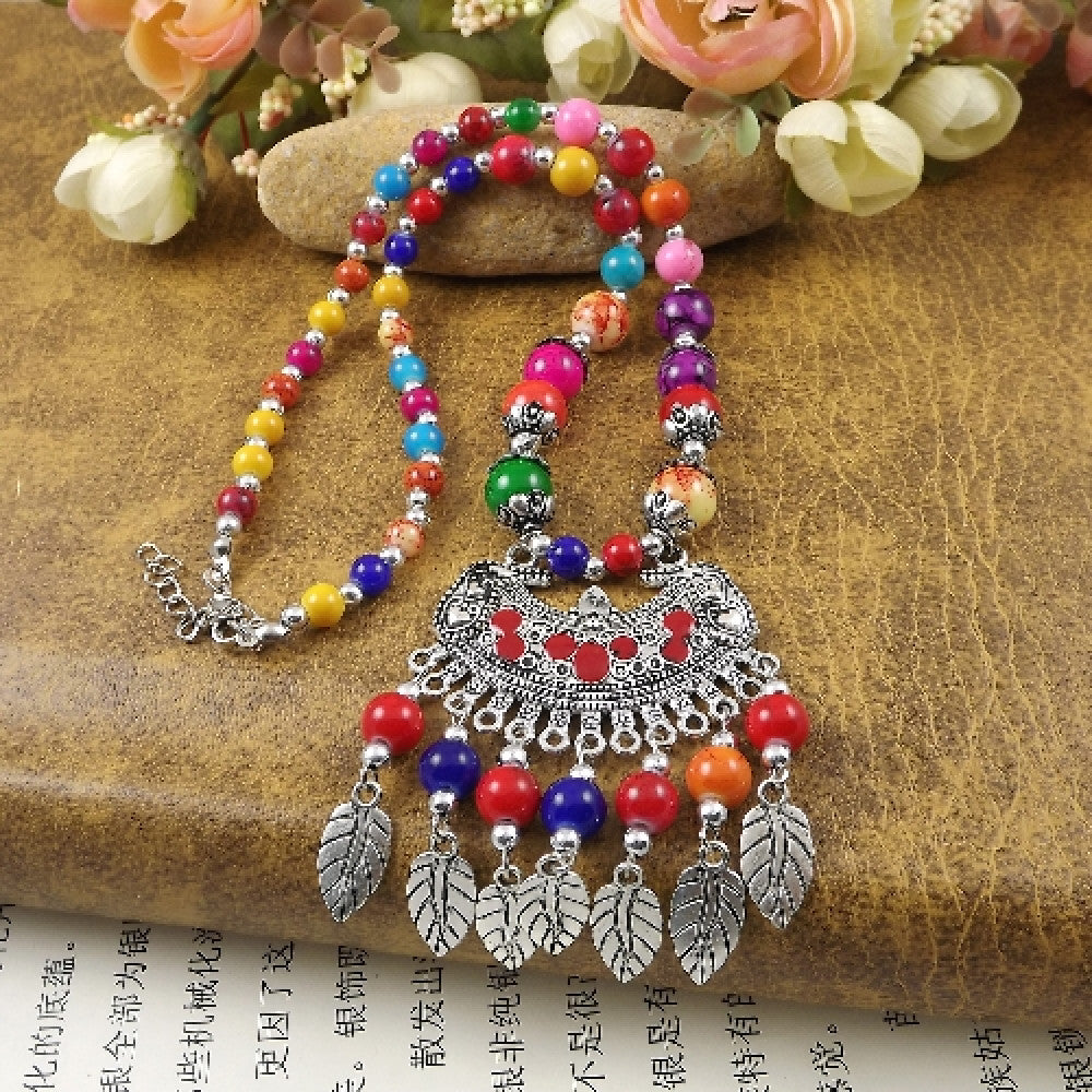 Vibrant colorful beaded necklace with intricate silver-toned leaf tassel pendant, featuring a mix of bright beads and vintage tribal design, ideal for bohemian fashion, showcased on a textured surface with floral accents
 01