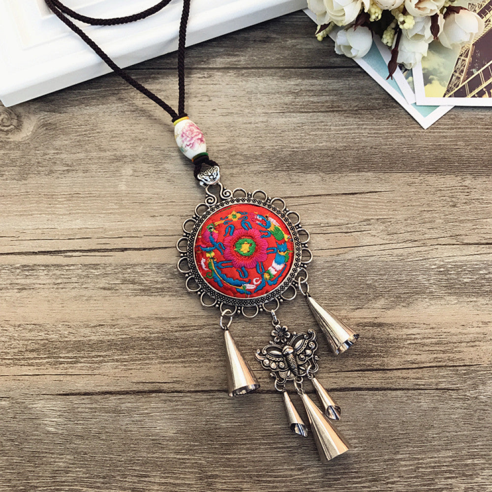 Vintage bohemian ethnic necklace with embroidered floral pendant, silver bells, and ceramic accent, displayed on a wooden surface with flowers, ideal for boho fashion enthusiasts 01