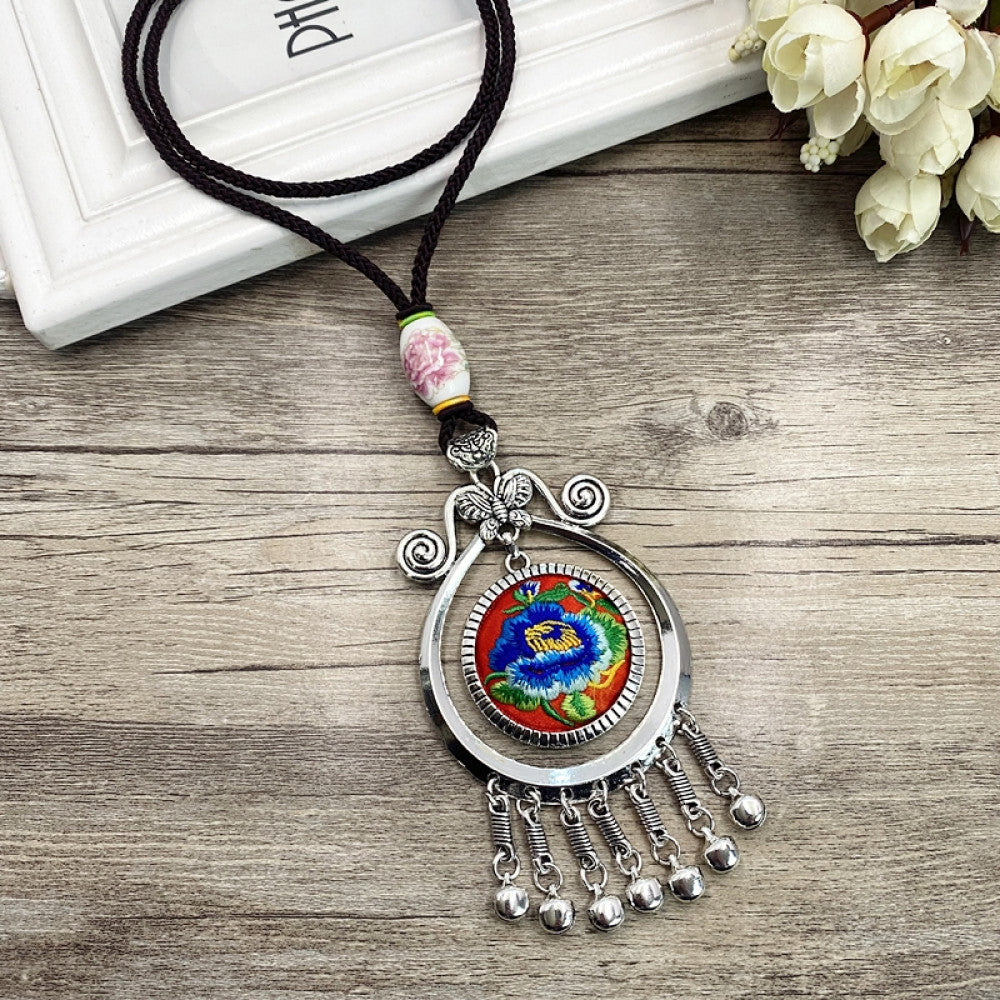 Vintage bohemian ethnic necklace with embroidered floral pendant, silver bells, and ceramic accent, displayed on a wooden surface with flowers, ideal for boho fashion enthusiasts 04