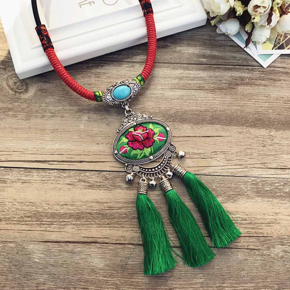 Vintage bohemian ethnic necklace with embroidered floral pendant, silver bells, and ceramic accent, displayed on a wooden surface with flowers, ideal for boho fashion enthusiasts 08