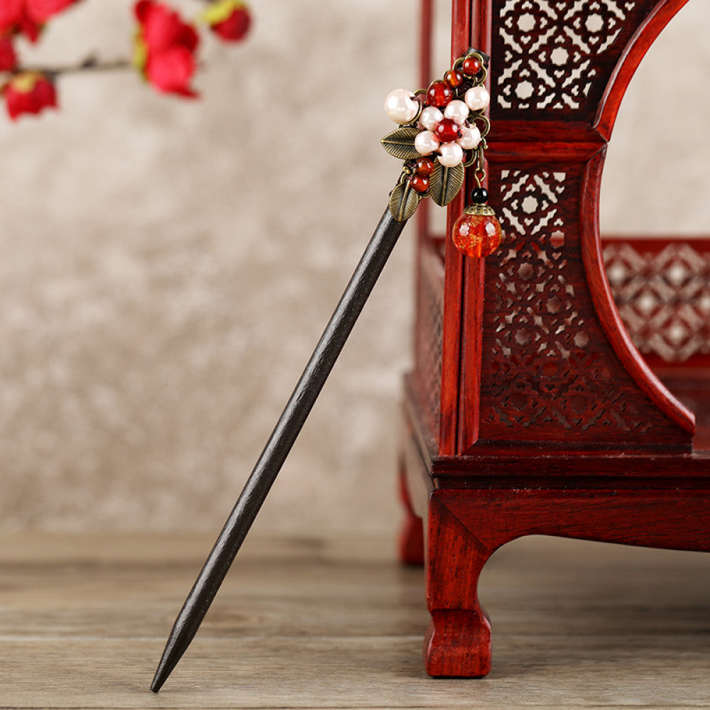 Boho vintage ethnic hair stick featuring a dark wooden-look handle, topped with light green flower, red bead, and green leaf decorative accents, displayed beside a red carved wooden decor