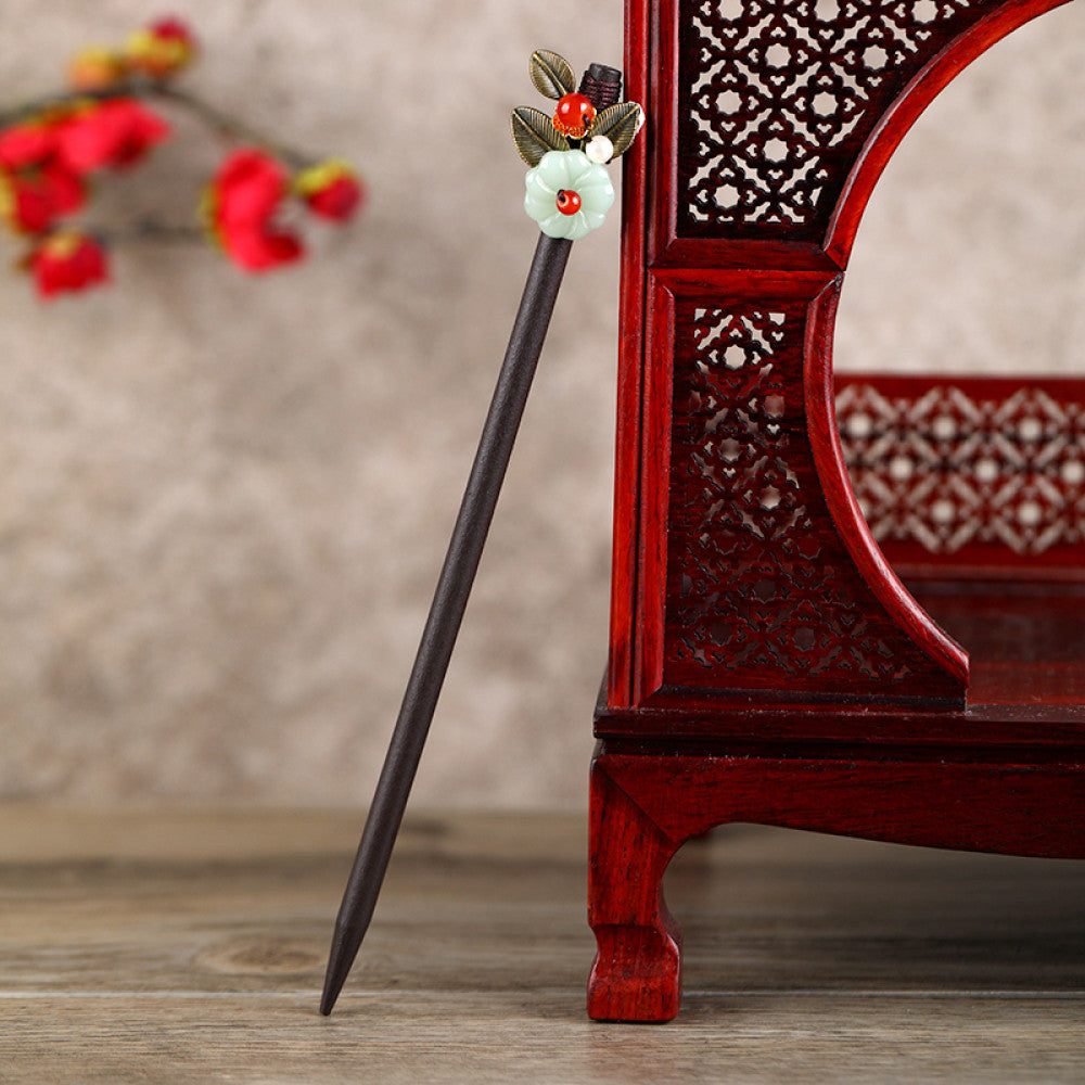 Boho vintage ethnic hair stick featuring a dark wooden-look handle, topped with light green flower, red bead, and green leaf decorative accents, displayed beside a red carved wooden decor (against a background with red florals), ideal for bohemian and traditional fashion enthusiasts in the US