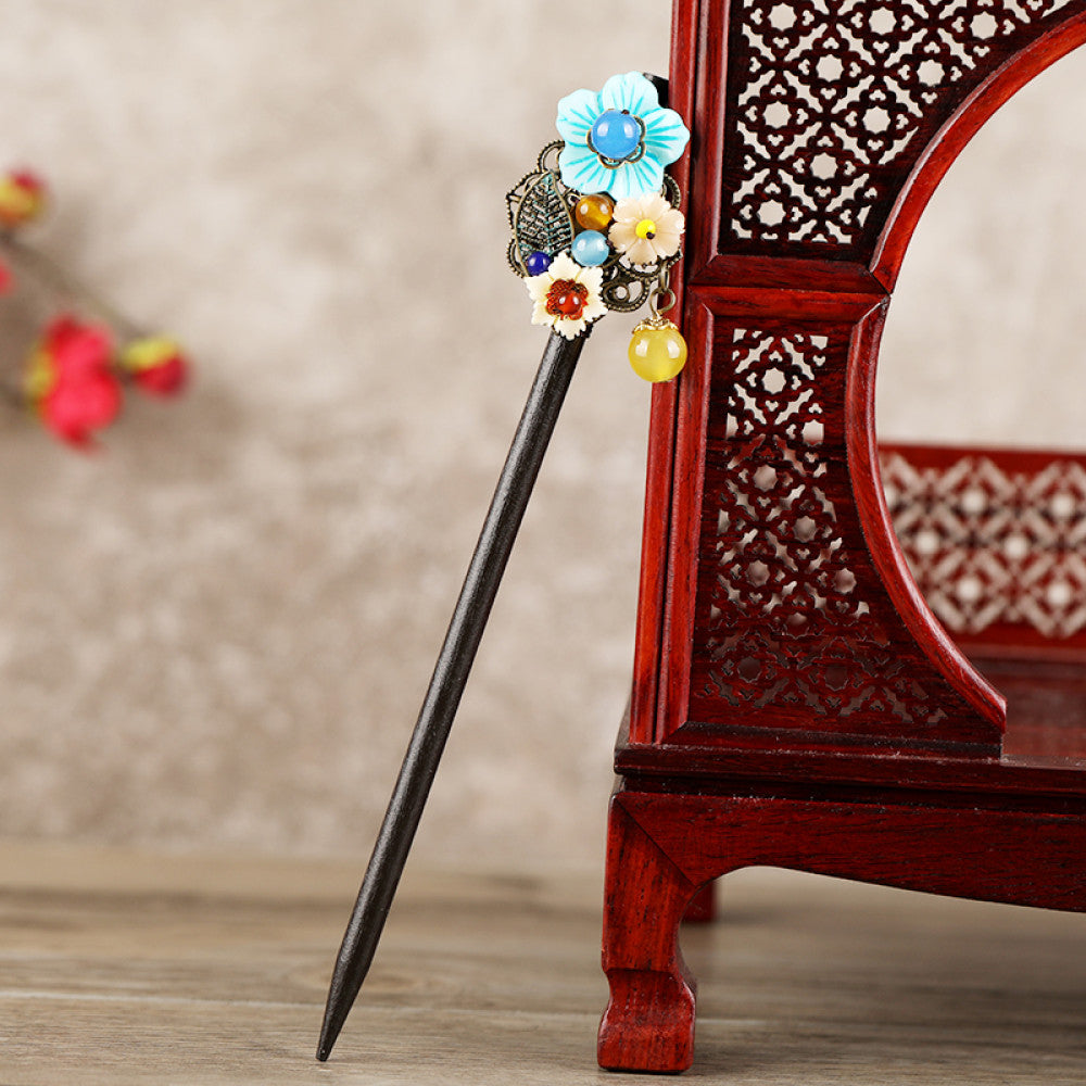 Boho vintage ethnic hair stick featuring a dark wooden-look handle, topped with light green flower, red bead, and green leaf decorative accents, displayed beside a red carved wooden decor , ideal for bohemian and traditional fashion enthusiasts in the US 03