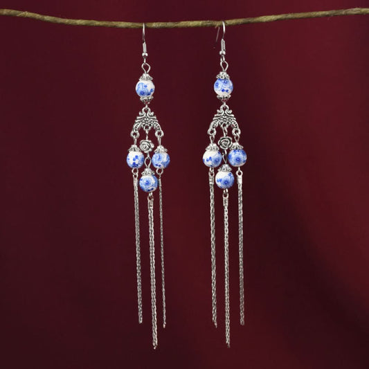 Pair of boho vintage ethnic silver-tone dangle earrings, featuring blue-and-white porcelain-style beads, intricate decorative details, and layered chain tassel fringe, hung on a twig against a rich red fabric background, ideal for bohemian and traditional fashion styling in the US