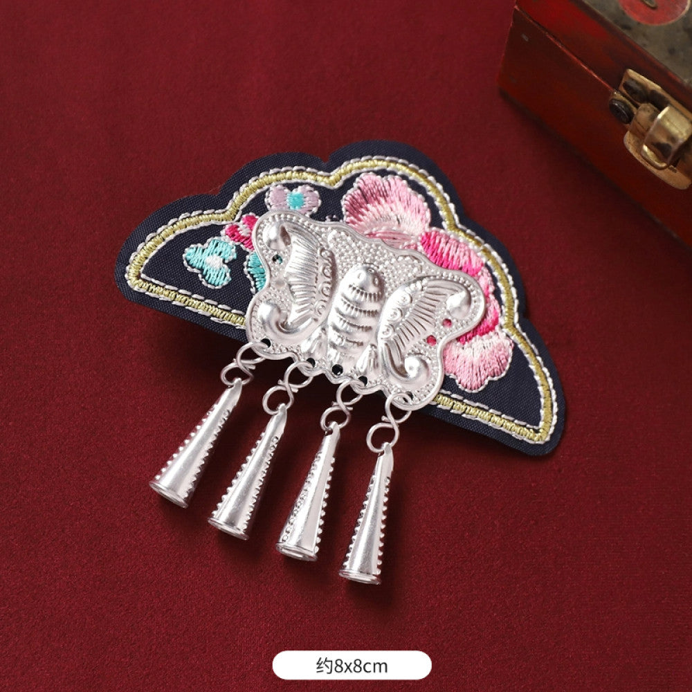 Boho vintage ethnic silver-tone decorative brooch featuring an intricate butterfly charm, pink floral embroidered accent (with blue details), and bell tassel pendants, displayed on a red fabric background