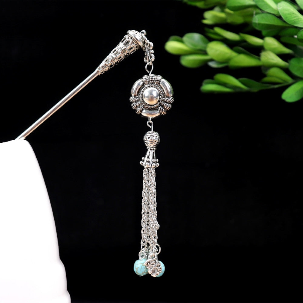 Boho vintage ethnic silver-tone hair stick featuring turquoise beads, an intricate round decorative element, and layered chain tassel fringe, displayed beside a white vessel green foliage 03