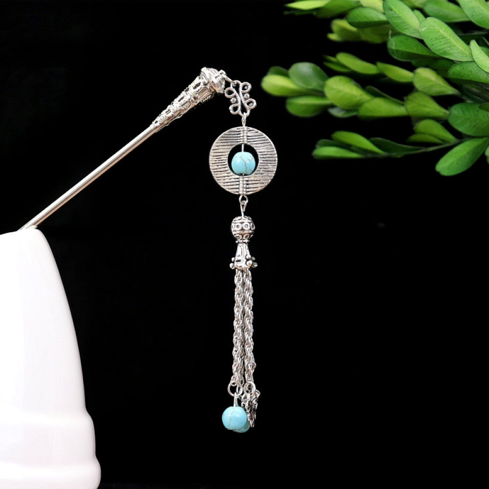 Boho vintage ethnic silver-tone hair stick featuring turquoise beads, an intricate round decorative element, and layered chain tassel fringe, displayed beside a white vessel green foliage , ideal for bohemian and traditional fashion enthusiasts in the US
