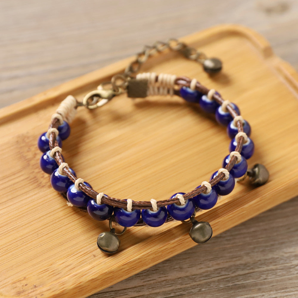 multicolored ceramic beads woven onto a brown leather cord, accented with small bronze bell charms. The adjustable design ensures a comfortable fit, adding a playful, earthy touch to any outfit.