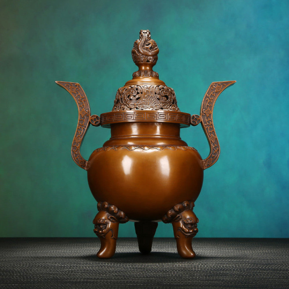 Intricate bronze Chinese incense burner with dragon-topped pierced lid, ornate curved handles, and lion-shaped feet, a Feng Shui symbol of wealth and elegance, displayed as a sophisticated home decor piece against a teal textured background