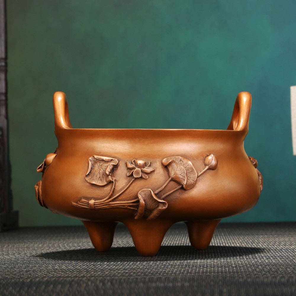 Intricate bronze Chinese incense burner with raised lotus flower carvings, curved handles, and three feet, a Feng Shui symbol of wealth and harmony, displayed as elegant home decor on a textured surface against a teal background