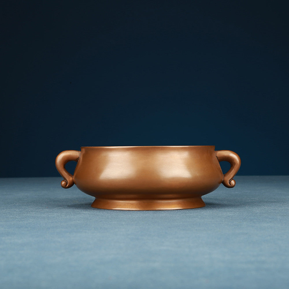 Polished bronze Chinese incense burner with elegant ruyi-shaped handles and a rounded body, a Feng Shui symbol of wealth and harmony, displayed as refined home decor against a dark blue backdrop on a textured surface