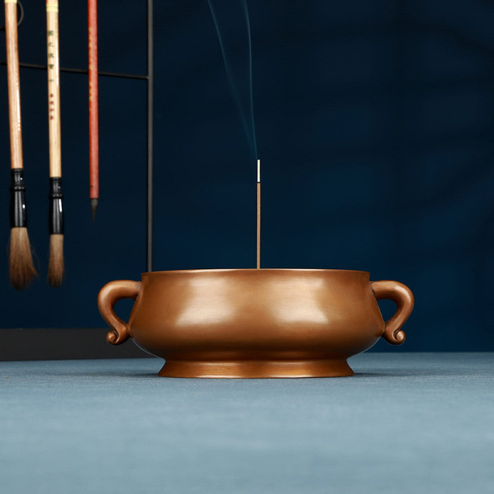 Polished bronze Chinese incense burner with elegant ruyi-shaped handles and a rounded body, a Feng Shui symbol of wealth and harmony, displayed as refined home decor against a dark blue backdrop on a textured surface 01