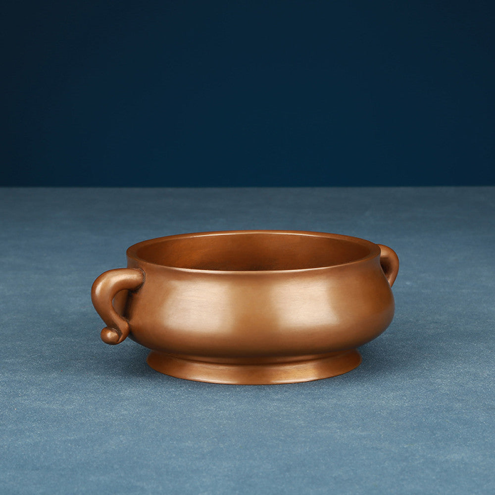 Polished bronze Chinese incense burner with elegant ruyi-shaped handles and a rounded body, a Feng Shui symbol of wealth and harmony, displayed as refined home decor against a dark blue backdrop on a textured surface 02