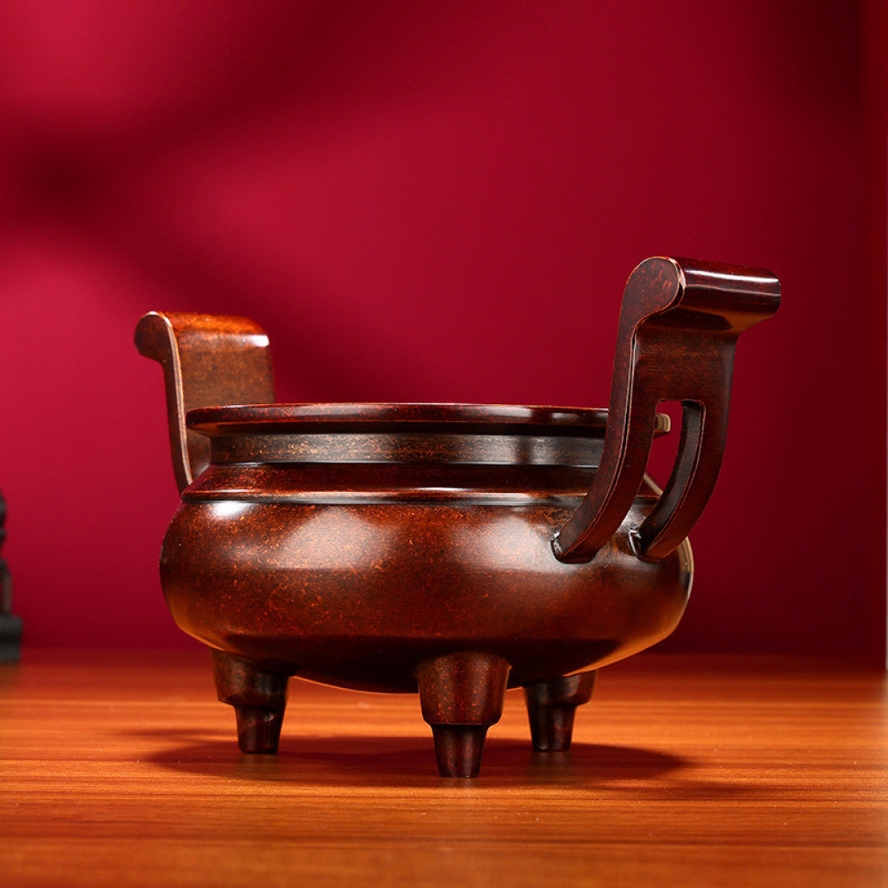 Intricate bronze Chinese incense burner with traditional curved handles and three feet, a Feng Shui symbol of wealth and harmony, displayed as elegant home decor on a wooden surface against a red background