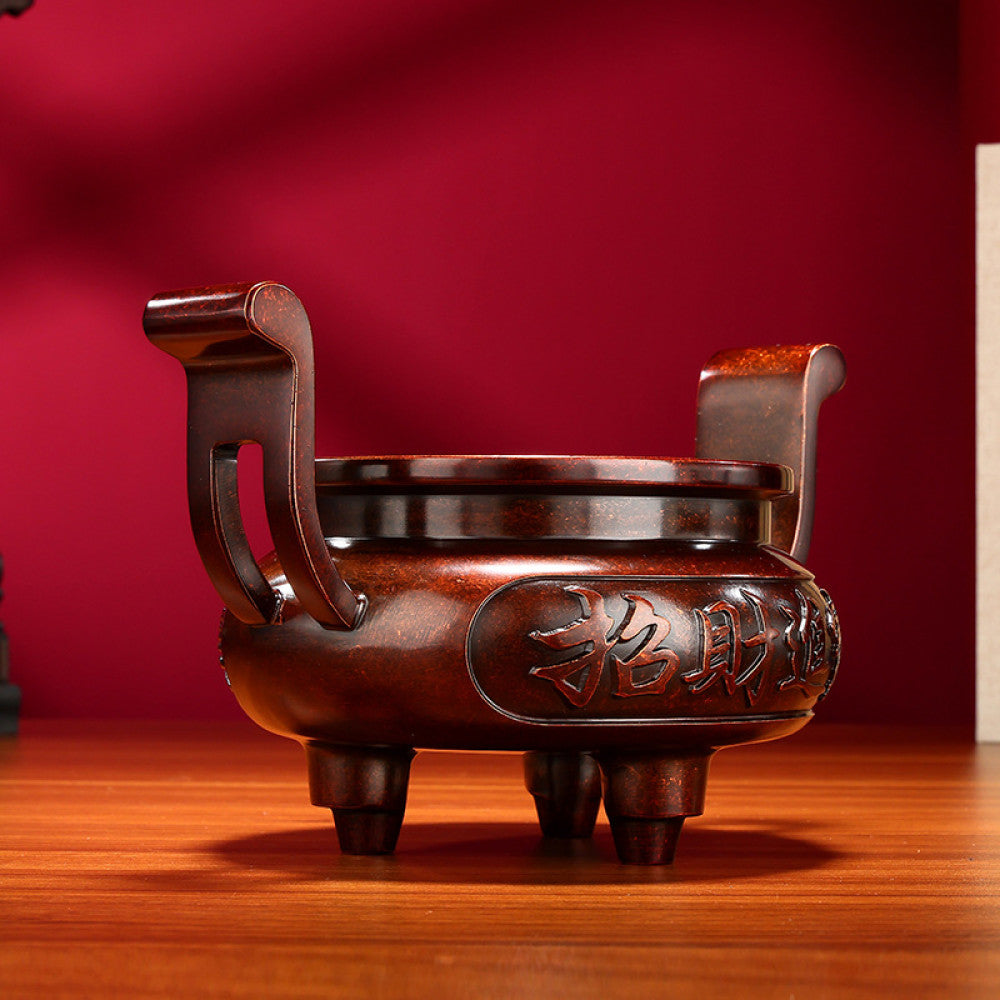 Intricate bronze Chinese incense burner with traditional curved handles, three feet, and "Wealth & Prosperity" carvings, a Feng Shui symbol of good fortune, displayed as elegant home decor on a wooden surface against a red background
