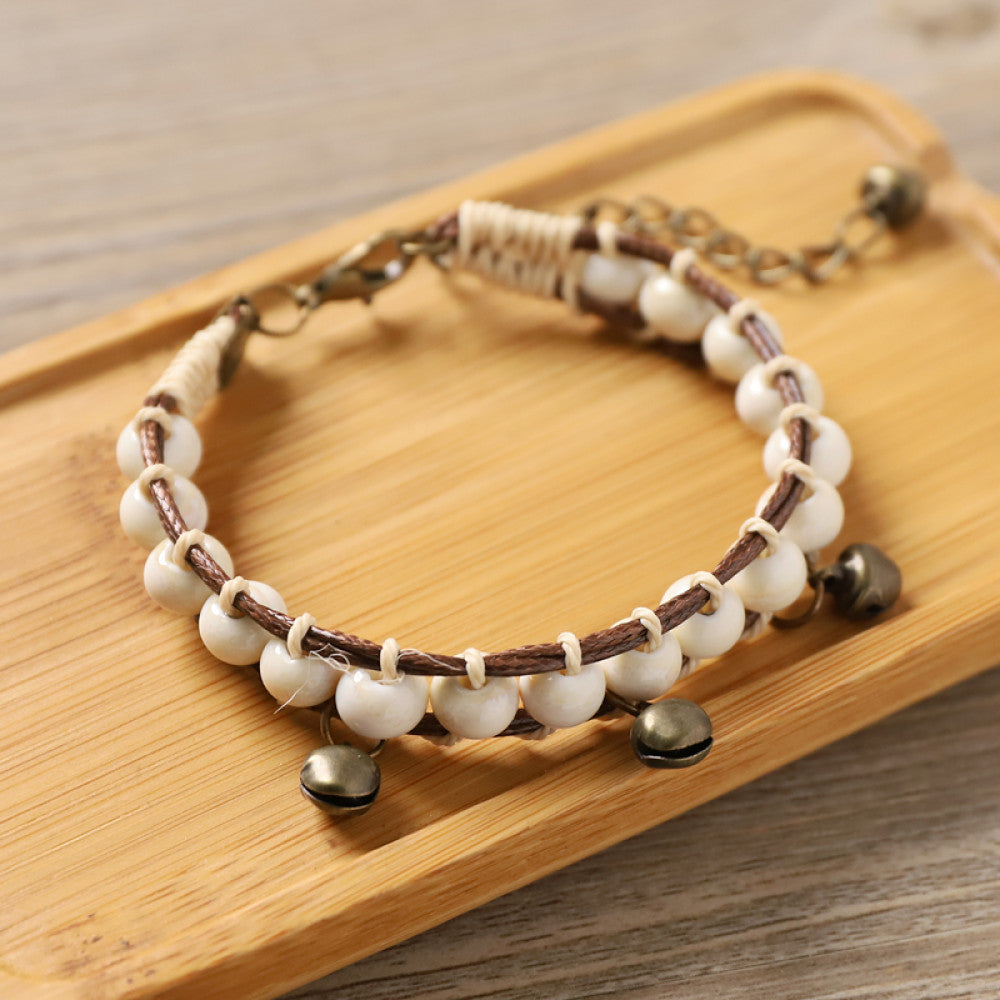 This bohemian bracelet features vibrant multicolored ceramic beads woven onto a brown leather cord, accented with small bronze bell charms