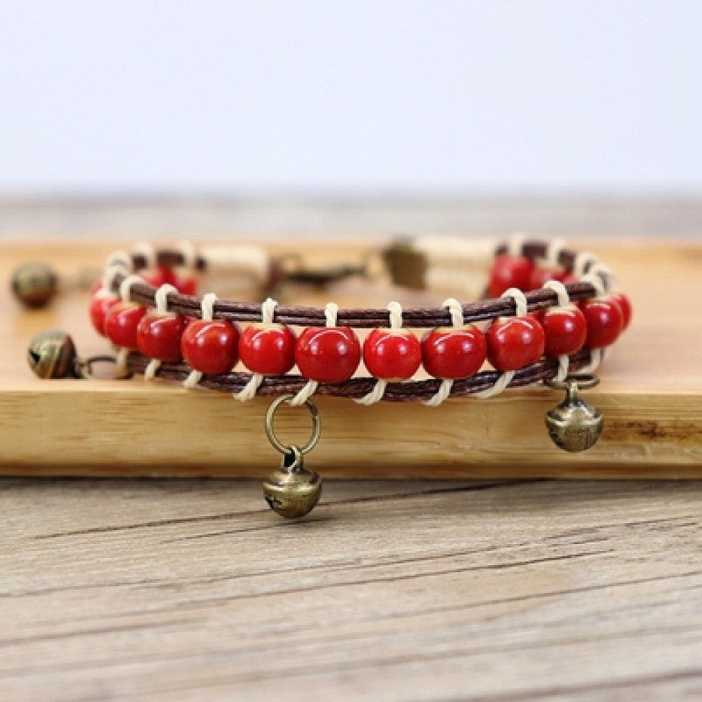 This bohemian bracelet features vibrant multicolored ceramic beads woven onto a brown leather cord, accented with small bronze bell charms. The adjustable design ensures a comfortable fit, adding a playful,