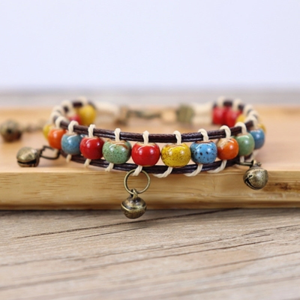 This bohemian bracelet features vibrant multicolored ceramic beads woven onto a brown leather cord, accented with small bronze bell charms. The adjustable design ensures a comfortable fit, adding a playful, earthy touch to any outfit