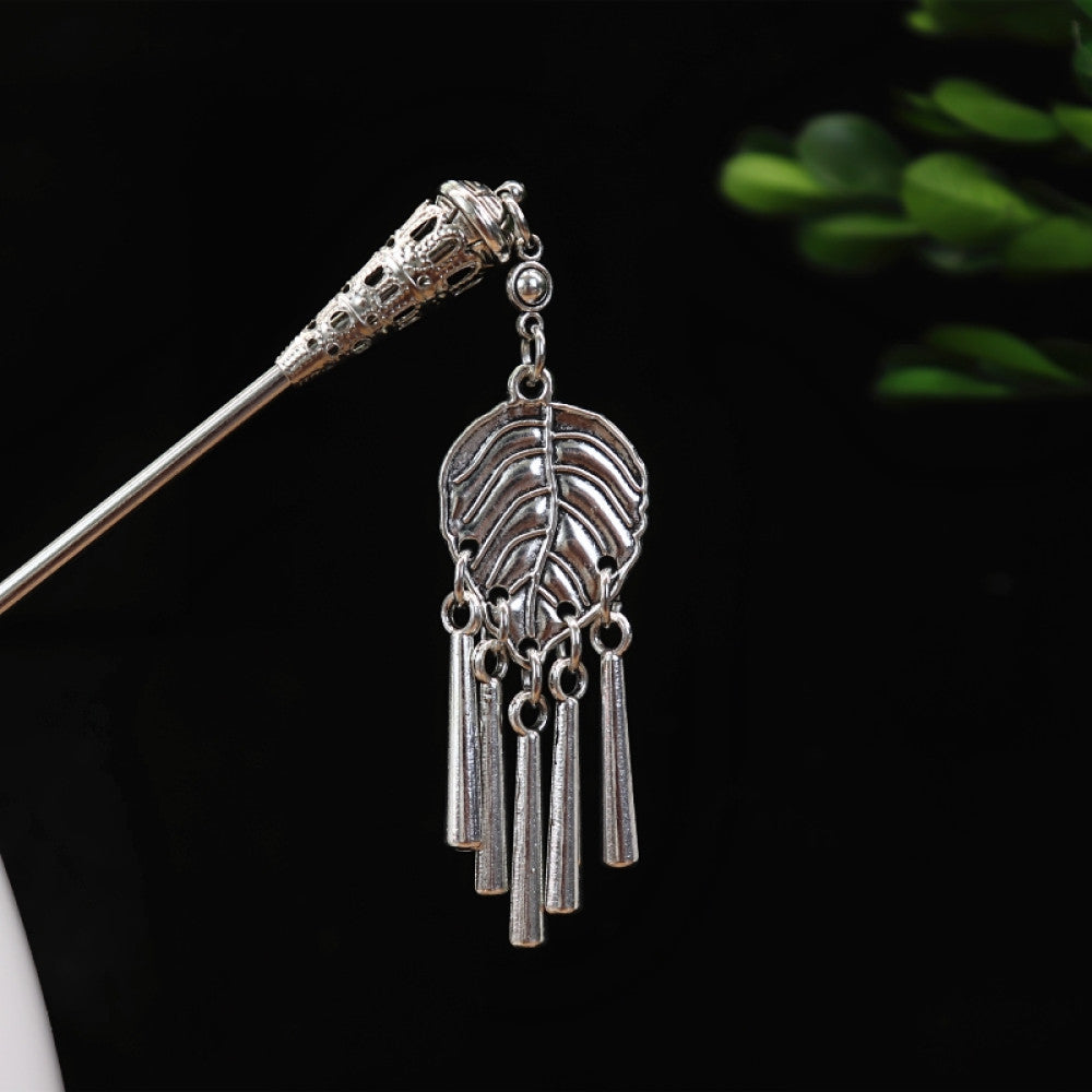 silver-tone hair stick featuring turquoise beads, an intricate round decorative element, and layered chain tassel fringe, displayed beside a white vessel green foliage