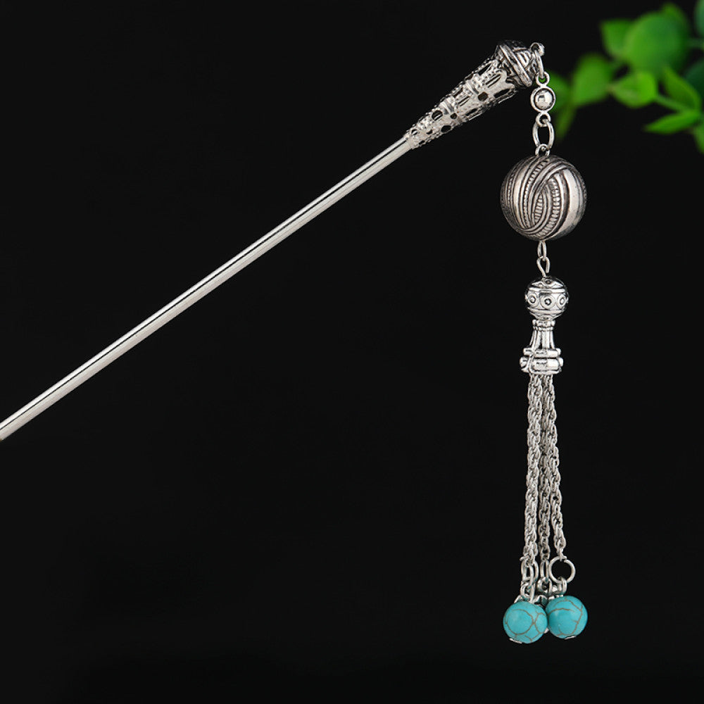 silver-tone hair stick featuring turquoise beads, an intricate round decorative element, and layered chain tassel fringe, displayed beside a white vessel green foliage 06