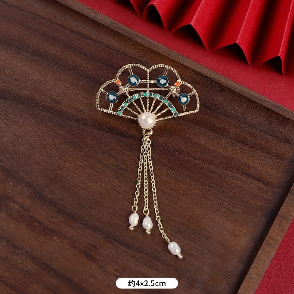 This elegant brooch features a delicate fan-shaped design, crafted with a gold-tone metal frame and adorned with vibrant multicolored gemstones. A lustrous pearl anchors the center, from which three graceful gold chains dangle, each finished with a small pearl drop. Perfect for adding a touch of sophistication to blazers, scarves, or special occasion outfits, this piece blends intricate craftsmanship with timeless, eye-catching charm.