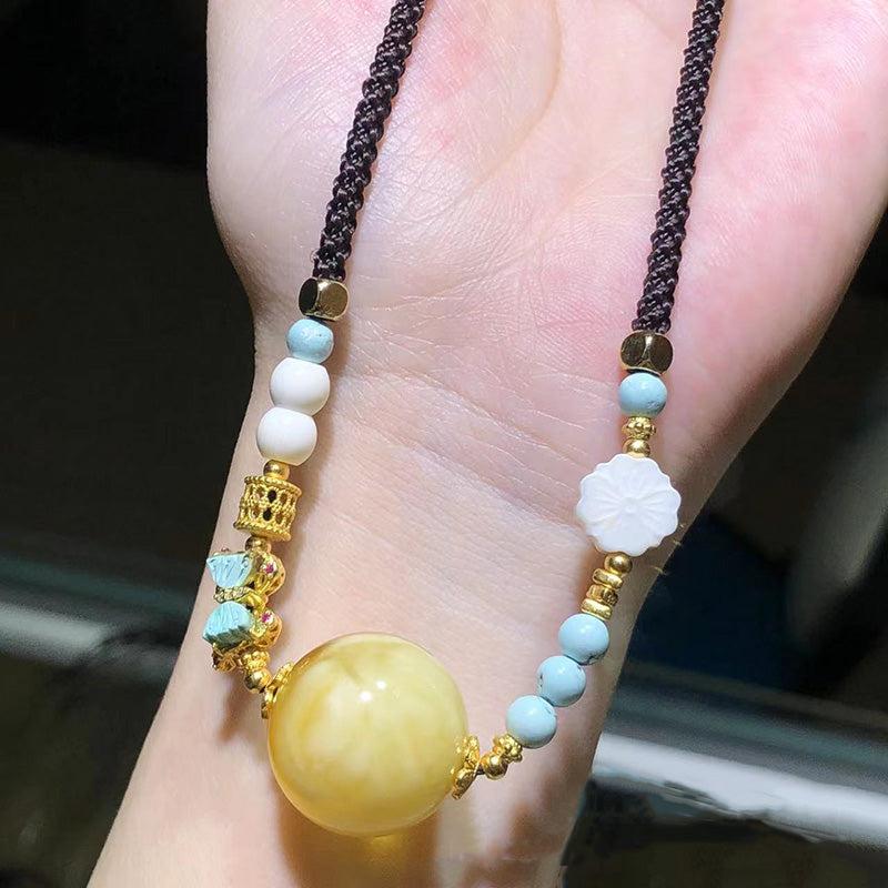 A stunning handmade necklace featuring a large, exquisite amber pendant paired with blue-green beads, white shell-shaped beads, and golden yellow decorative elements, all strung on a dark woven cord.