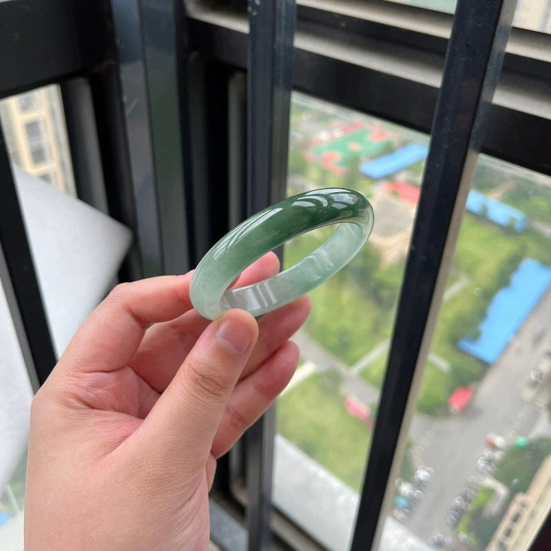 Hand holding icy green natural jade bangle against city view through window, Chinese jade jewelry