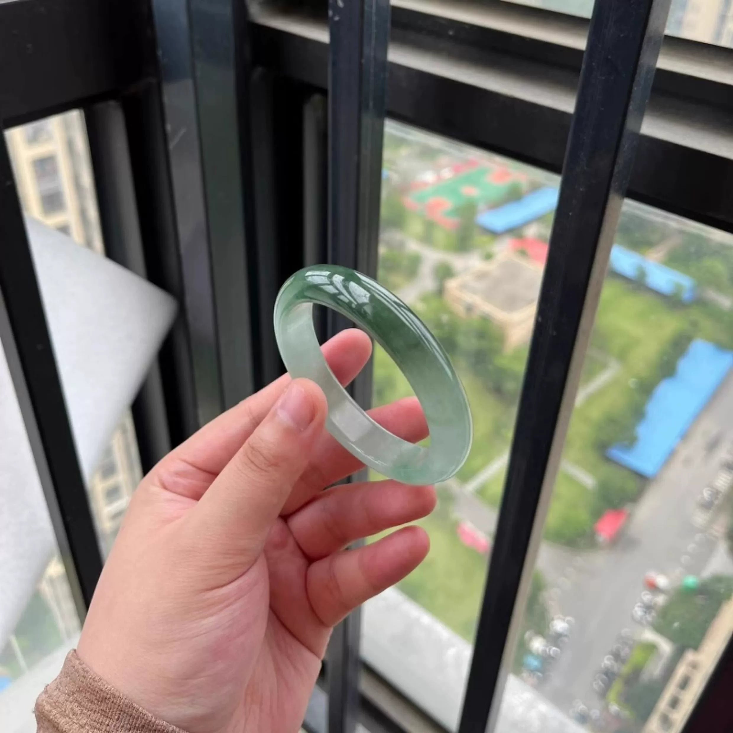Hand holding icy green natural jade bangle against city view through window, Chinese jade jewelry 01