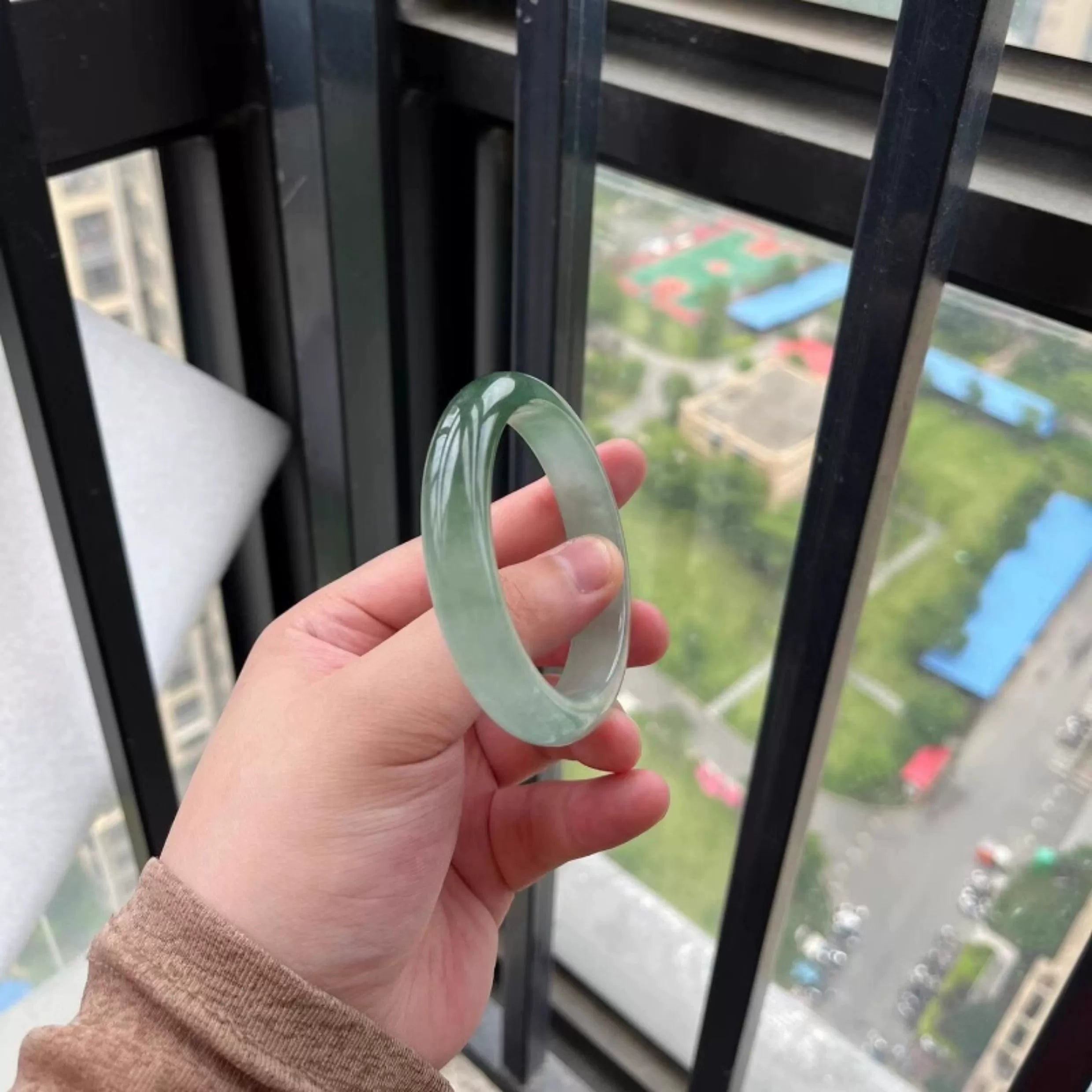 Hand holding icy green natural jade bangle against city view through window, Chinese jade jewelry 03