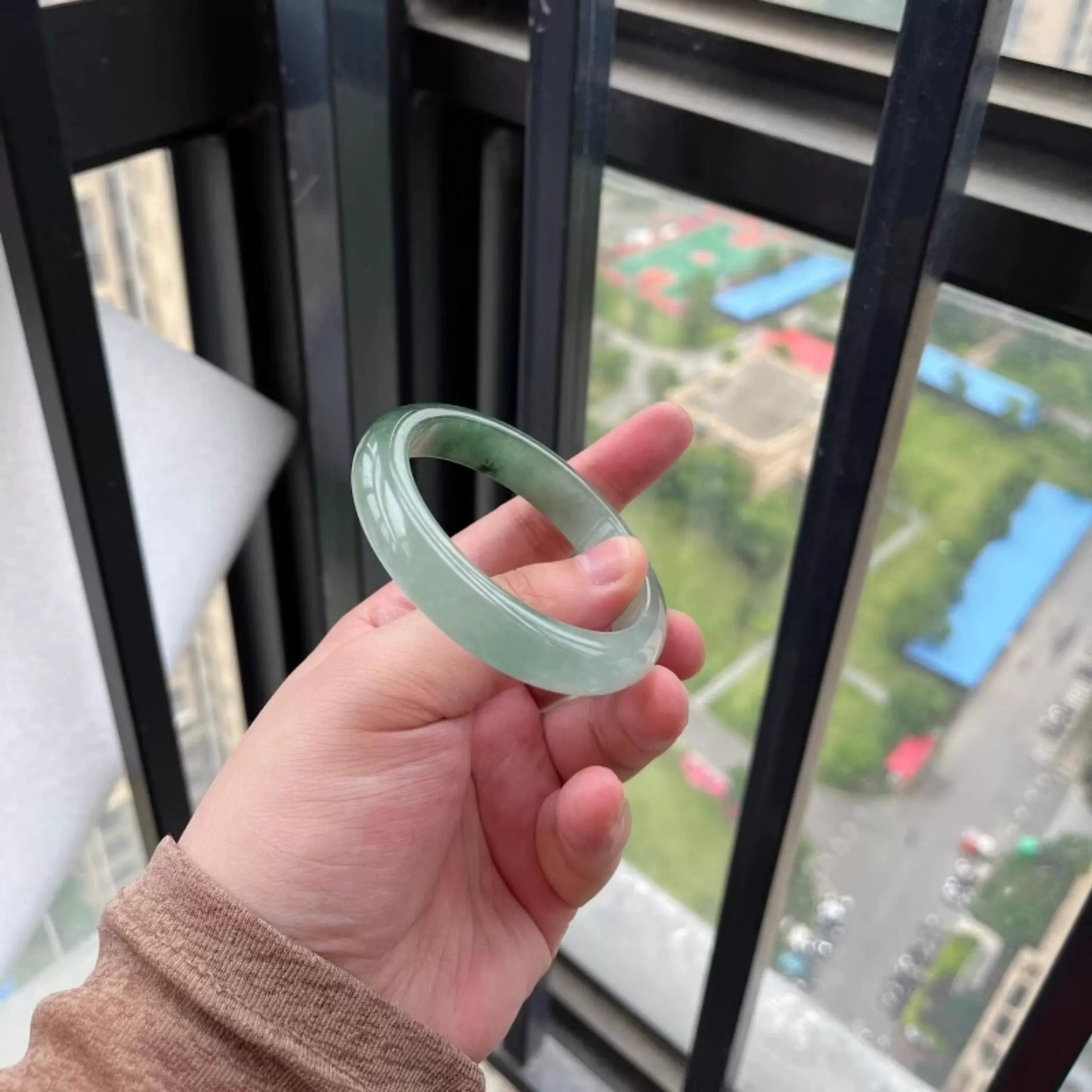 Hand holding icy green natural jade bangle against city view through window, Chinese jade jewelry 04