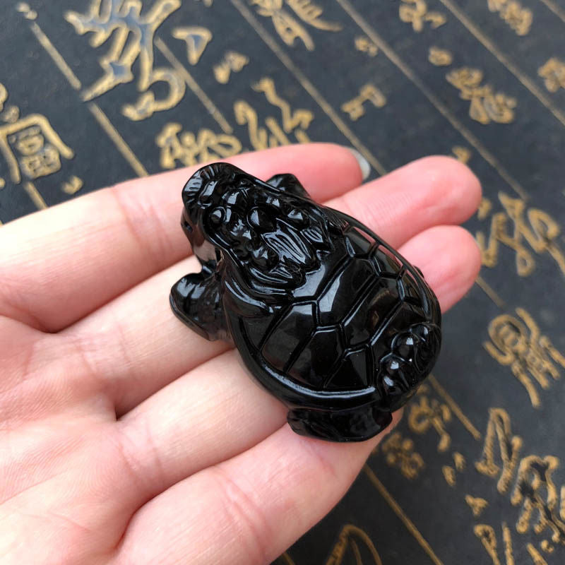 A hand - held natural black obsidian amulet carved into the shape of a dragon - turtle, showcasing fine craftsmanship and a glossy finish.