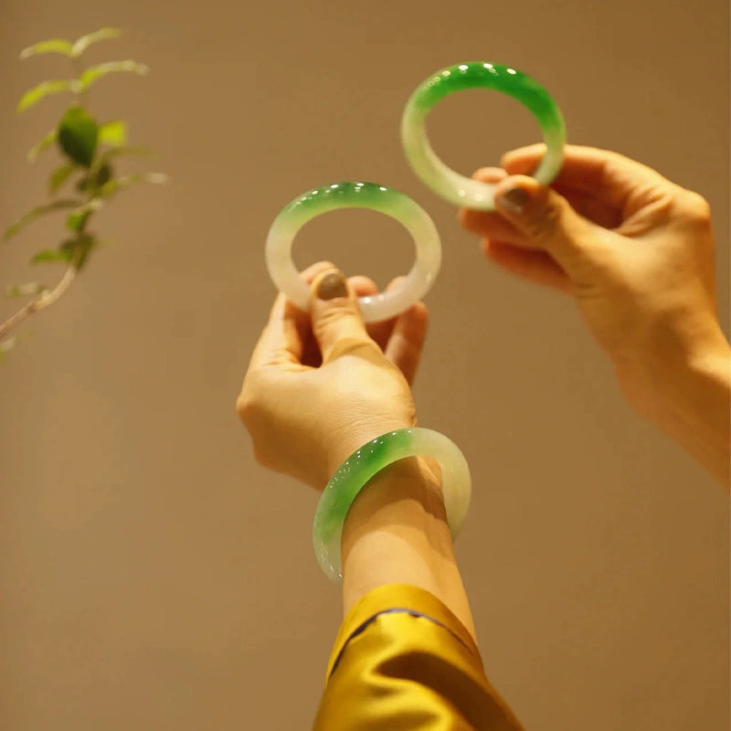 Hands holding multiple natural icy green gradient jadeite bangles with plant background