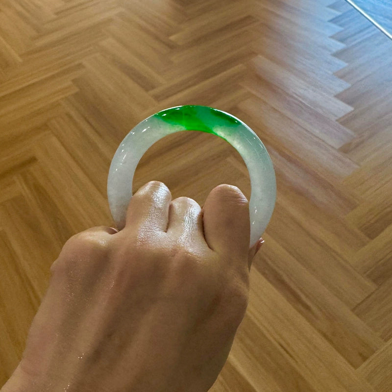 Hand holding natural icy jadeite bangle with vivid green patch on white base against herringbone wood floor