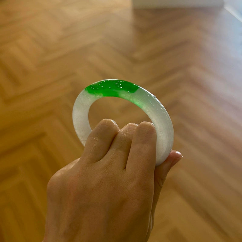 Hand holding natural icy jadeite bangle with vivid green patch on white base against herringbone wood floor 01