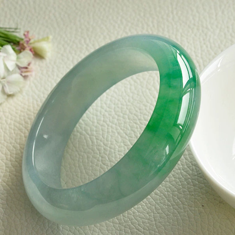 Natural icy jadeite bangle with vivid green patches placed near white dish and flowers on textured surface