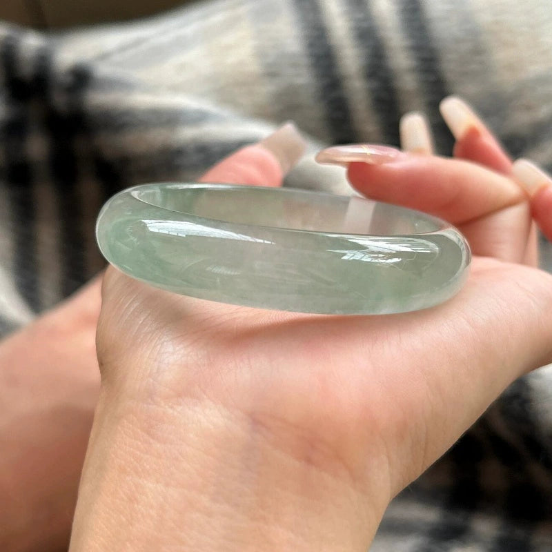 Hand holding natural icy light green jade bangle against plaid fabric