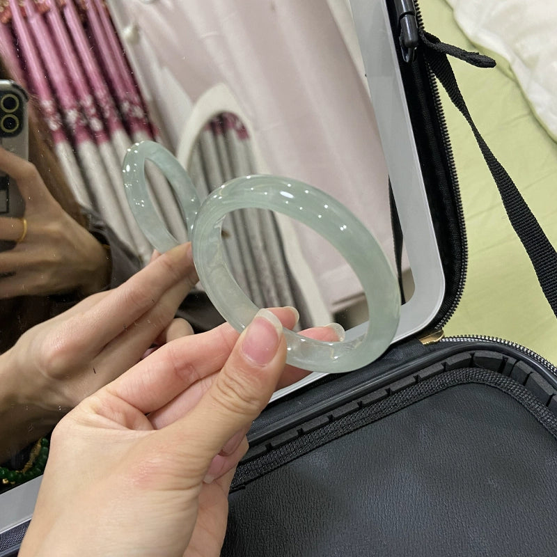 Hands holding two natural icy light green jade bangles by jewelry case