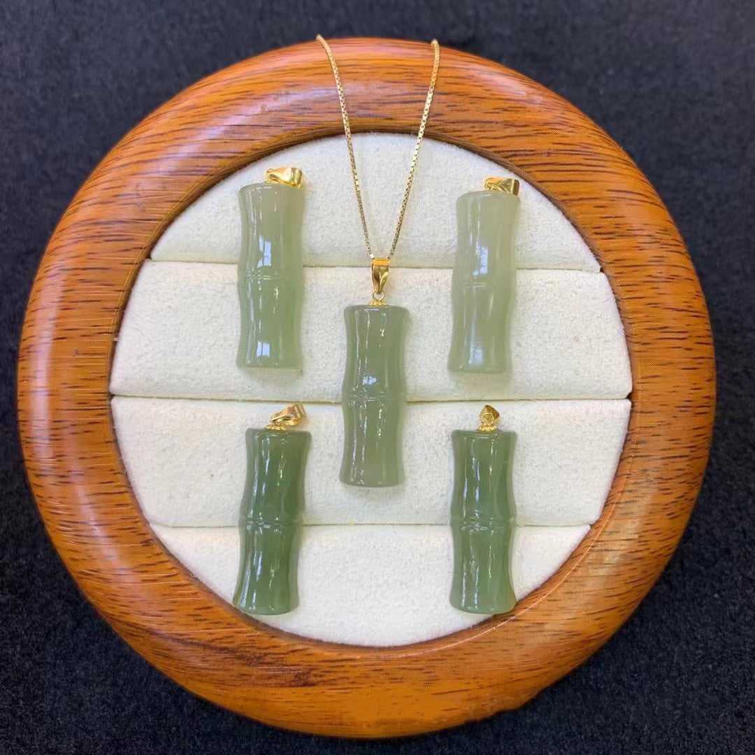 Set of natural nephrite jade bamboo - shaped pendants, each with gold - plated attachments, displayed on a round wooden jewelry tray with a soft, white - cushioned interior. One pendant is on a gold - toned chain, while the others are placed separately.