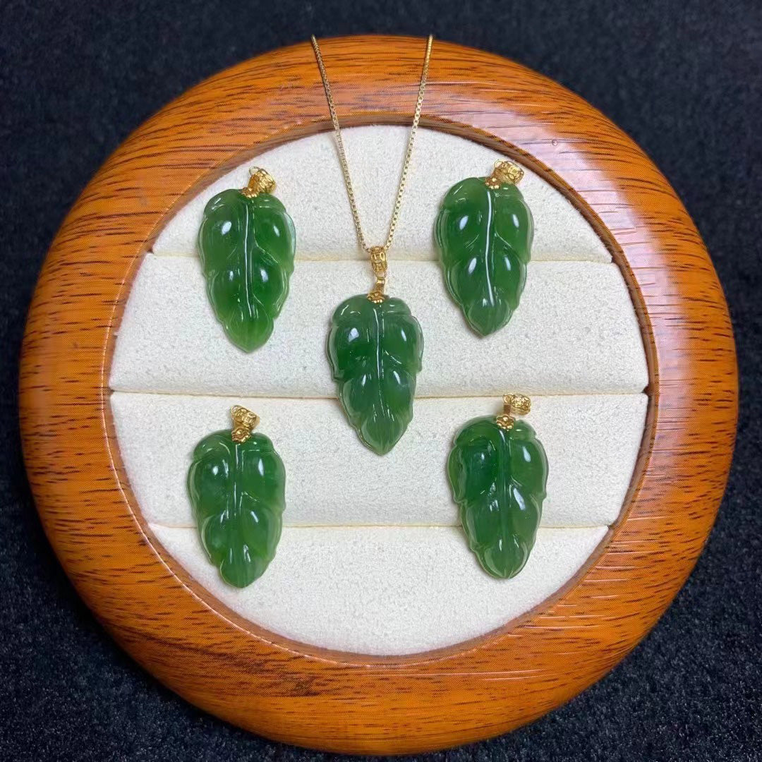 Set of natural nephrite jade pendants carved into leaf shapes, each adorned with gold - plated attachments. One pendant is suspended from a gold - toned chain, while the rest are placed separately on a round wooden jewelry tray with a soft, white - cushioned interior.
