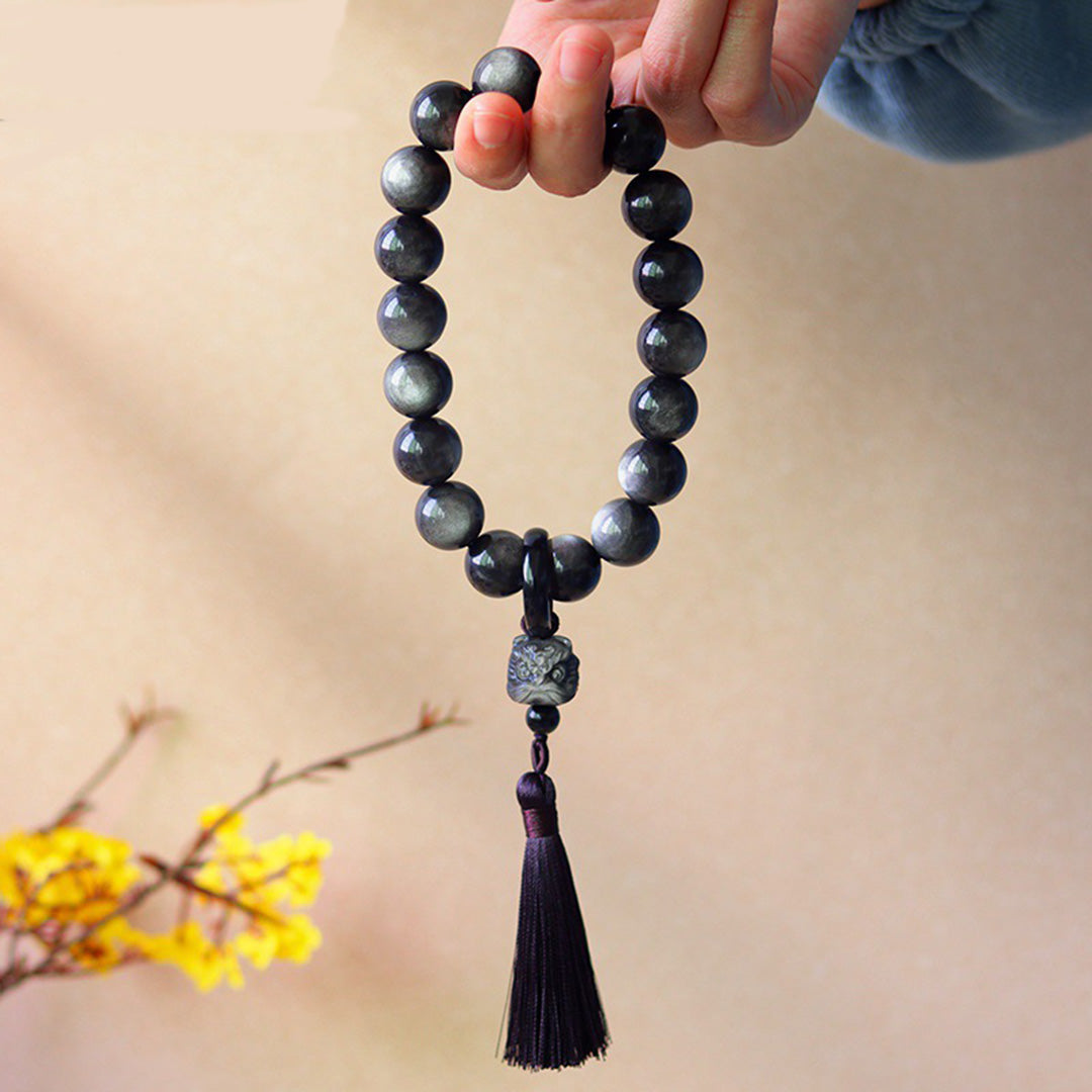 A hand - held natural rainbow obsidian bead bracelet featuring dark beads with subtle iridescent sheens, accompanied by a pendant attached to the bracelet.