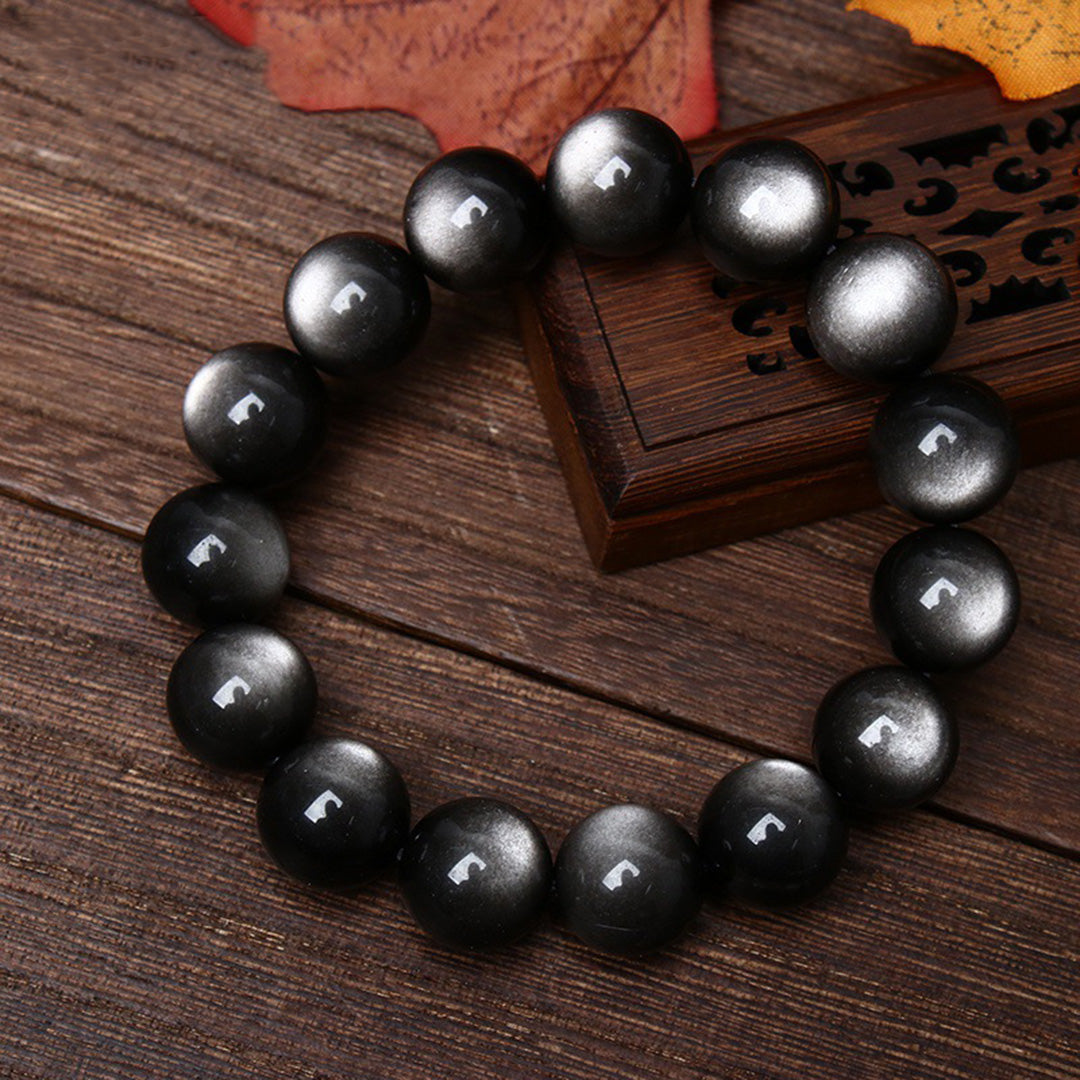 A natural rainbow obsidian bead bracelet with a distinct silver - sheen effect, placed on a wooden surface alongside a carved wooden box and a fallen autumn leaf 01