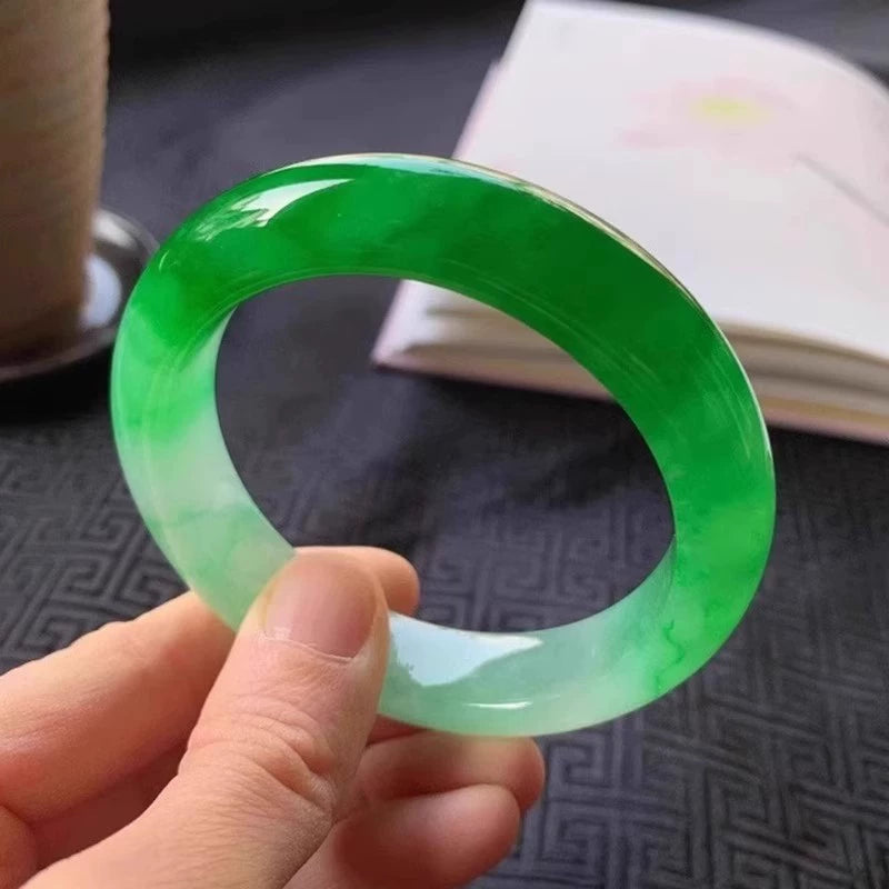 Hand holding natural vivid green gradient jadeite bangle over traditional patterned fabric with open book in background