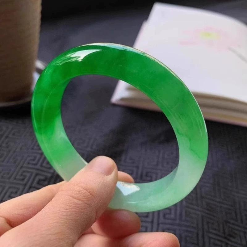 Hand holding natural vivid green gradient jadeite bangle over traditional patterned fabric with open book in background 01