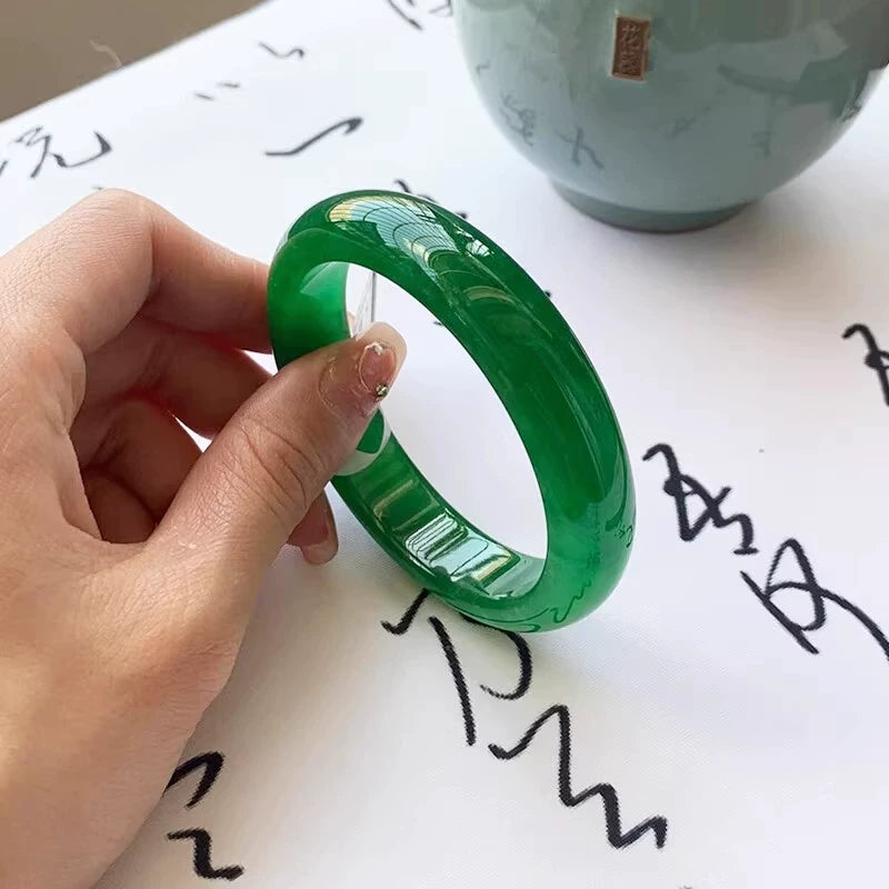 Hand holding natural vivid green gradient jadeite bangle over traditional patterned fabric with open book in background